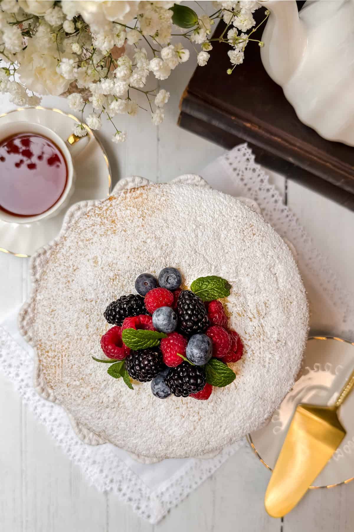 Round Irish Tea Cake dusted with powdered sugar and topped with blackberries, raspberries, blueberries, and mint leaves on a lace-adorned plate. Beside it, a cup of tea, a gold cake serving knife, and white flowers are visible on a wooden table.