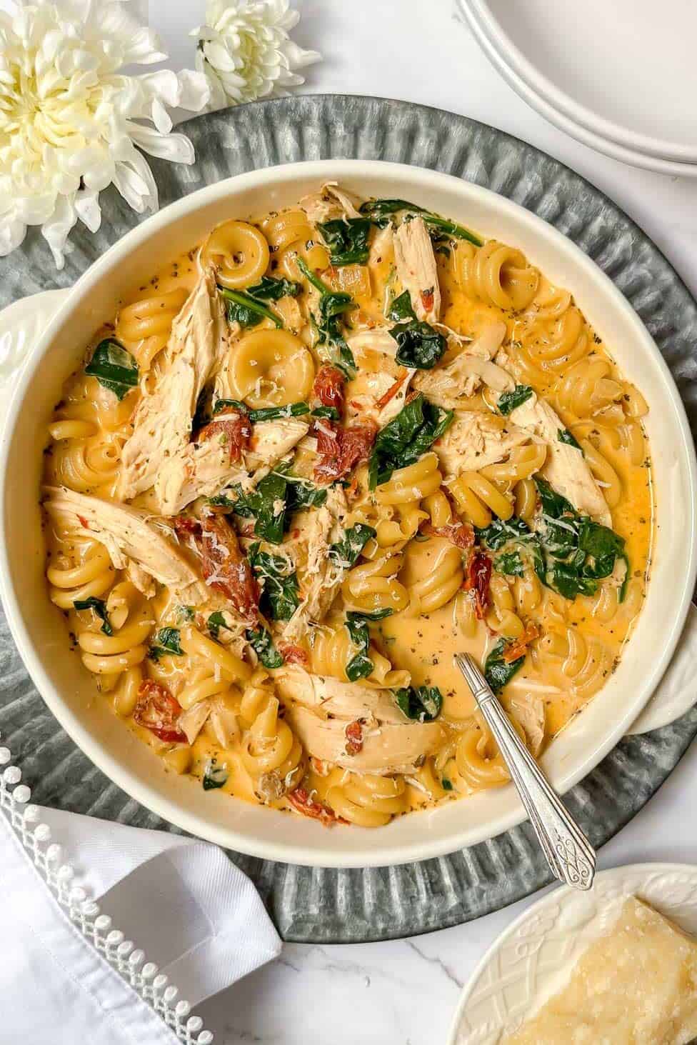 A bowl of creamy pasta with spinach, shredded chicken, and sun-dried tomatoes sits on a gray platter, reminiscent of a cozy "marry me" chicken soup. A grater with cheese is nearby. White flowers and a folded napkin embellish the setting, crafting an inviting dining atmosphere.