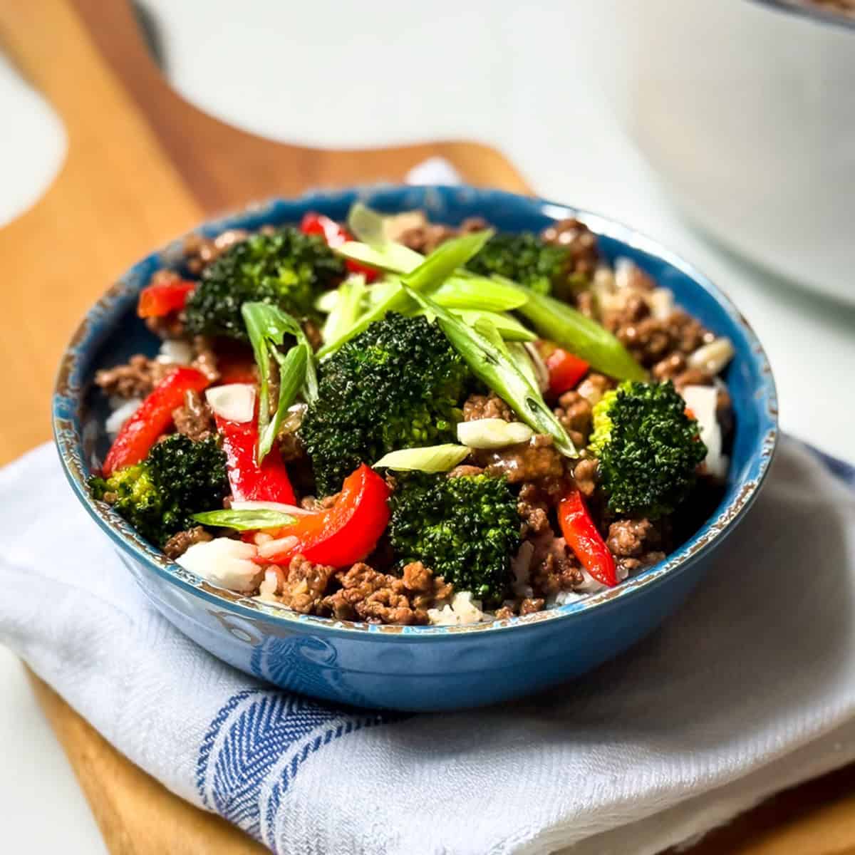 A blue bowl filled with savory Beef and Broccoli, red bell peppers, and sliced green onions served over white rice, placed on a folded white and blue cloth napkin.