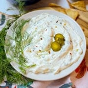 A plate of creamy dill pickle dip garnished with two green olives, fresh dill, and red pepper flakes, with pita chips in the background.