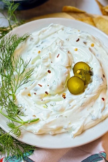 A plate of creamy dill pickle dip garnished with two green olives, fresh dill, and red pepper flakes, with pita chips in the background.
