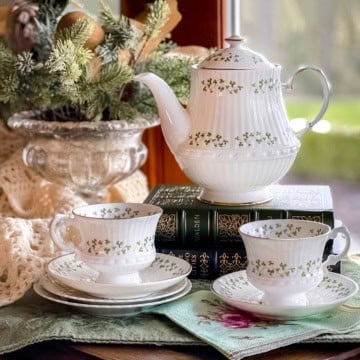 White shamrock teapot and matching teacups with green floral patterns evoke an Irish Afternoon Tea scene, displayed on stacked books beside a window with a leafy plant in the background.