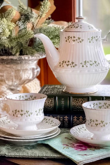 White shamrock teapot and matching teacups with green floral patterns evoke an Irish Afternoon Tea scene, displayed on stacked books beside a window with a leafy plant in the background.