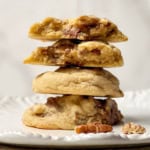 A stack of three thick cookies, the top two split open to reveal a gooey praline caramel and pecan filling, sits on a white plate with a few pecan pieces beside them.
