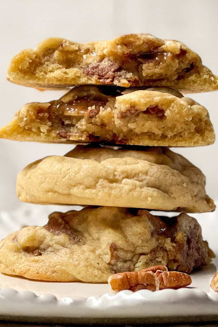 A stack of three thick cookies, the top two split open to reveal a gooey praline caramel and pecan filling, sits on a white plate with a few pecan pieces beside them.