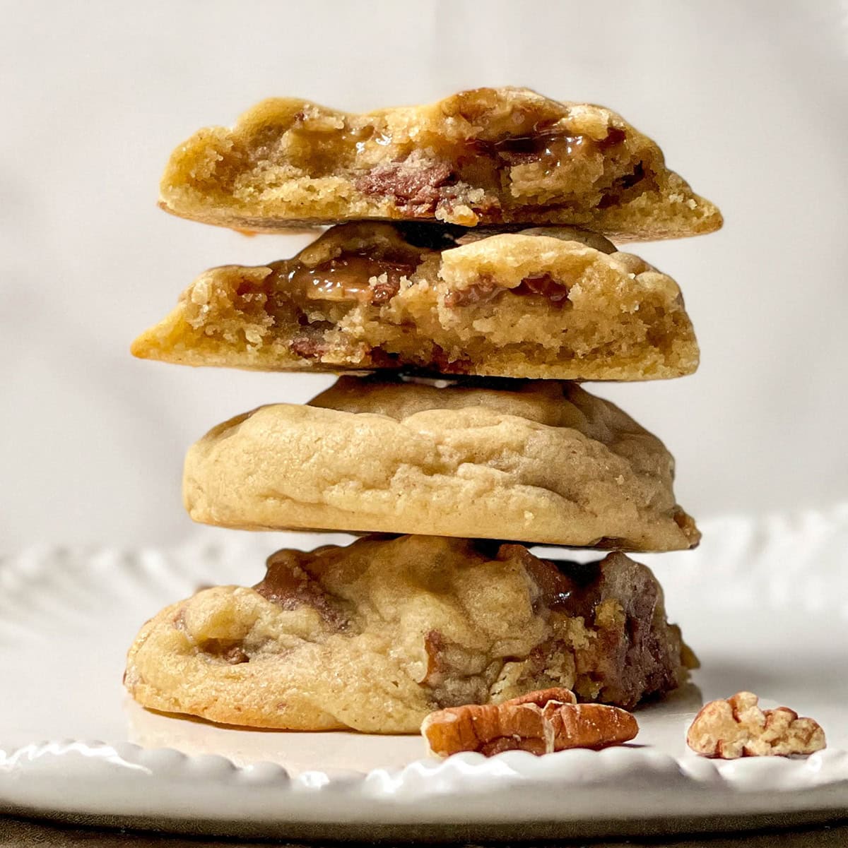 A stack of three thick cookies, the top two split open to reveal a gooey praline caramel and pecan filling, sits on a white plate with a few pecan pieces beside them.