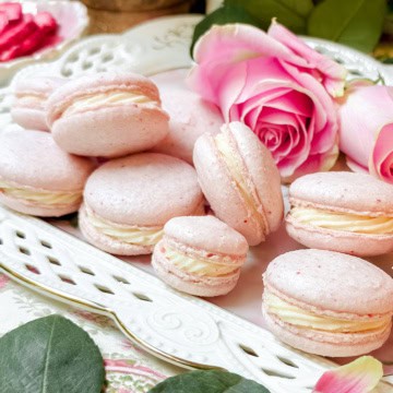 A plate of pink macarons with cream filling, made from a delightful macaron recipe, is displayed alongside a pink rose on a decorative white tray.