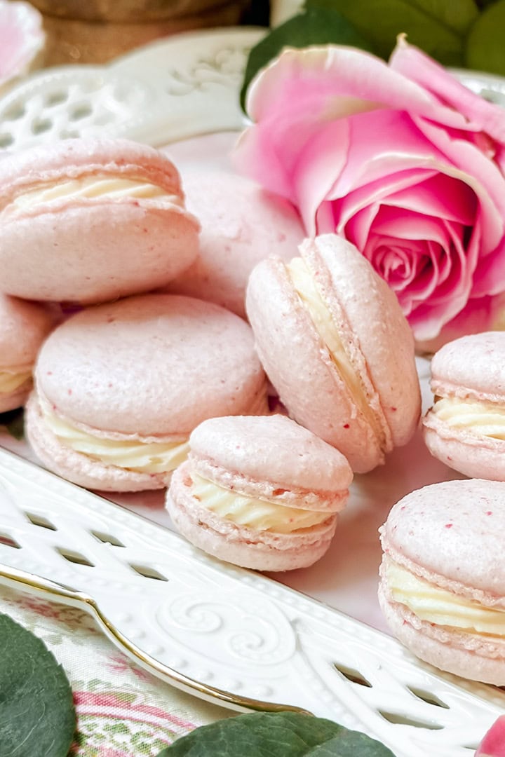A plate of pink macarons with cream filling, made from a delightful macaron recipe, is displayed alongside a pink rose on a decorative white tray.