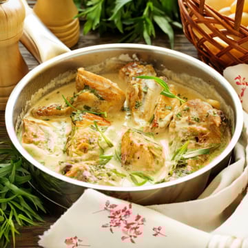 Cooked chicken pieces in a creamy sauce, inspired by Abraham Lincoln's Chicken Fricassee, are garnished with fresh herbs in a metal pan, set on a table with a patterned napkin, fresh greens, and a bread basket nearby.