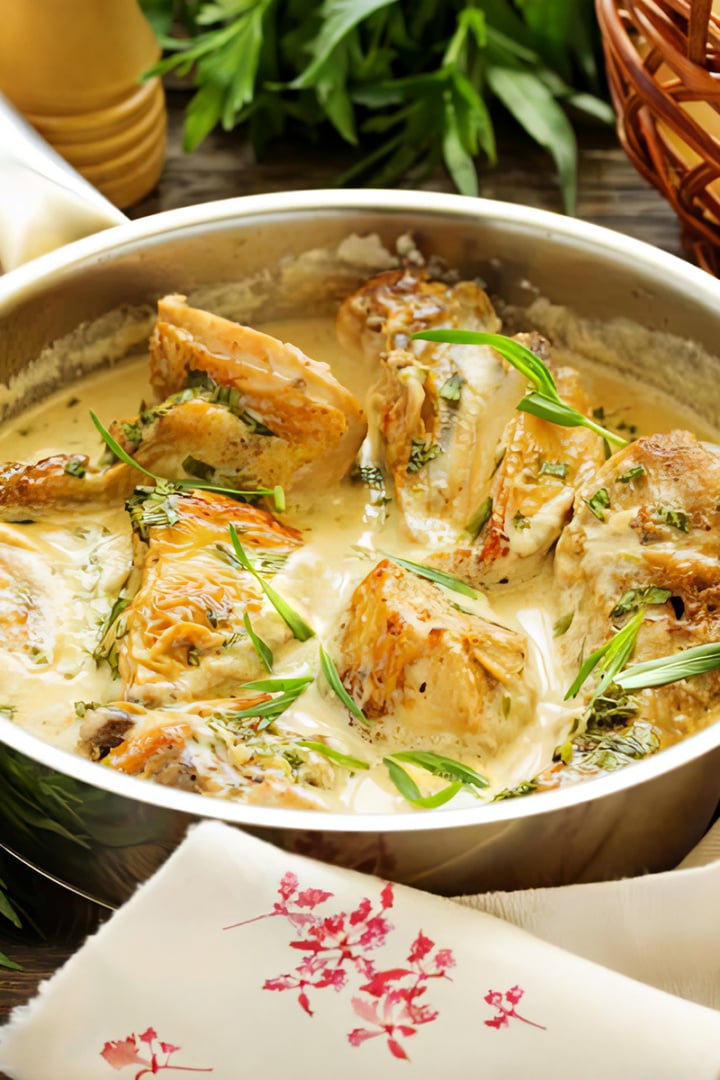 Cooked chicken pieces in a creamy sauce, inspired by Abraham Lincoln's Chicken Fricassee, are garnished with fresh herbs in a metal pan, set on a table with a patterned napkin, fresh greens, and a bread basket nearby.