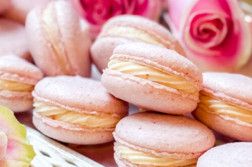 A plate of pink macarons with cream filling stacked on a floral napkin, hinting at a delightful macaron recipe. Pink roses grace the background, adding a romantic touch to the scene.