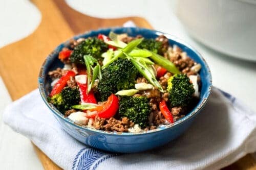 A blue bowl brimming with flavorful Beef and Broccoli, enhanced by vibrant red bell peppers. Garnished with sliced green onions, the dish rests on a white cloth adorned with blue stripes atop a rustic wooden board.