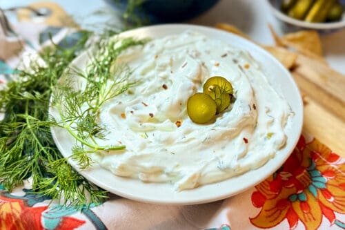 A bowl of creamy white dip garnished with sliced green pickles and fresh dill, placed on a floral-patterned cloth. Thin dill sprigs are scattered around, enhancing the presentation.