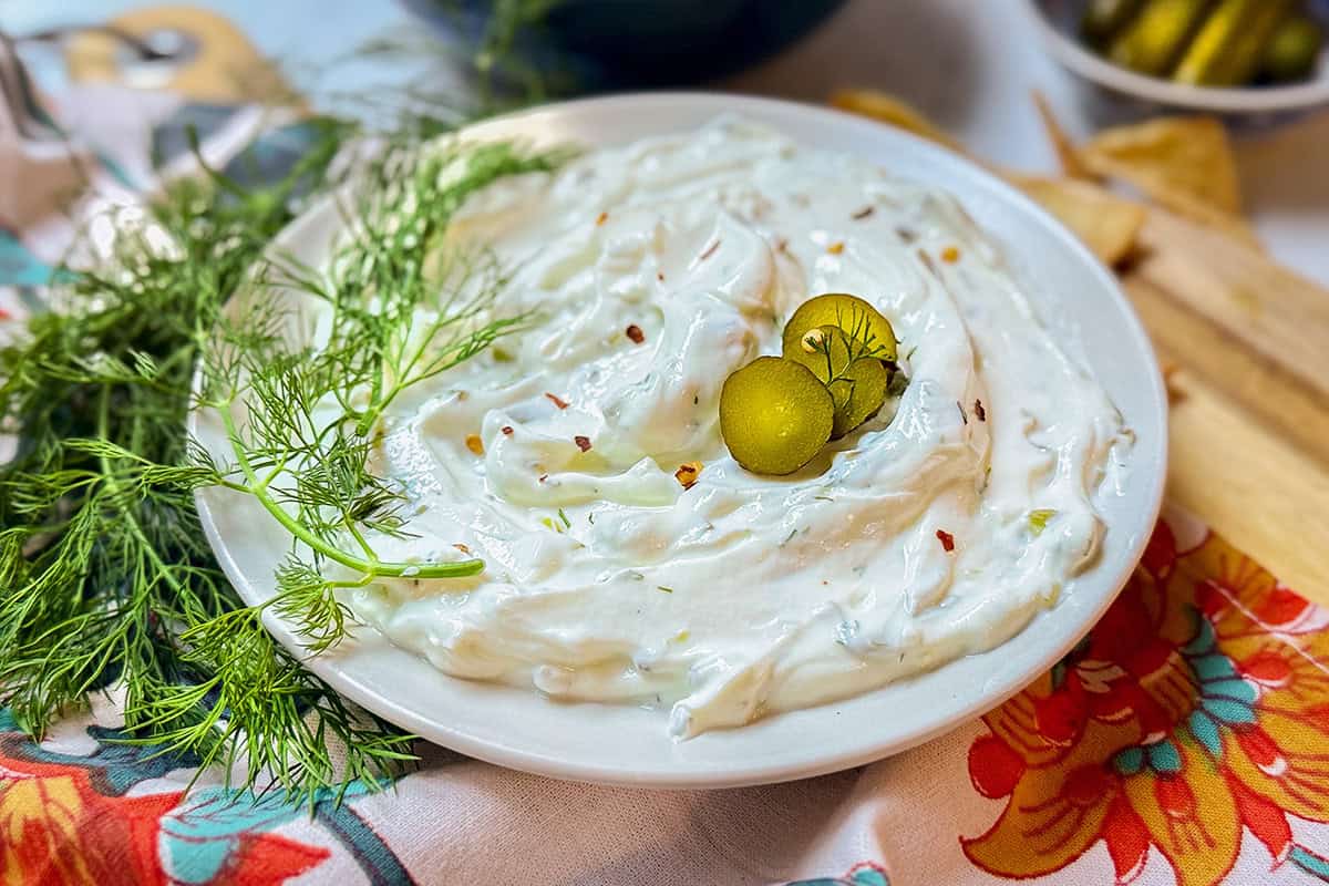 A plate of creamy dill pickle dip garnished with two pickle slices and fresh dill, set on a floral-patterned cloth with extra dill sprigs beside the plate.