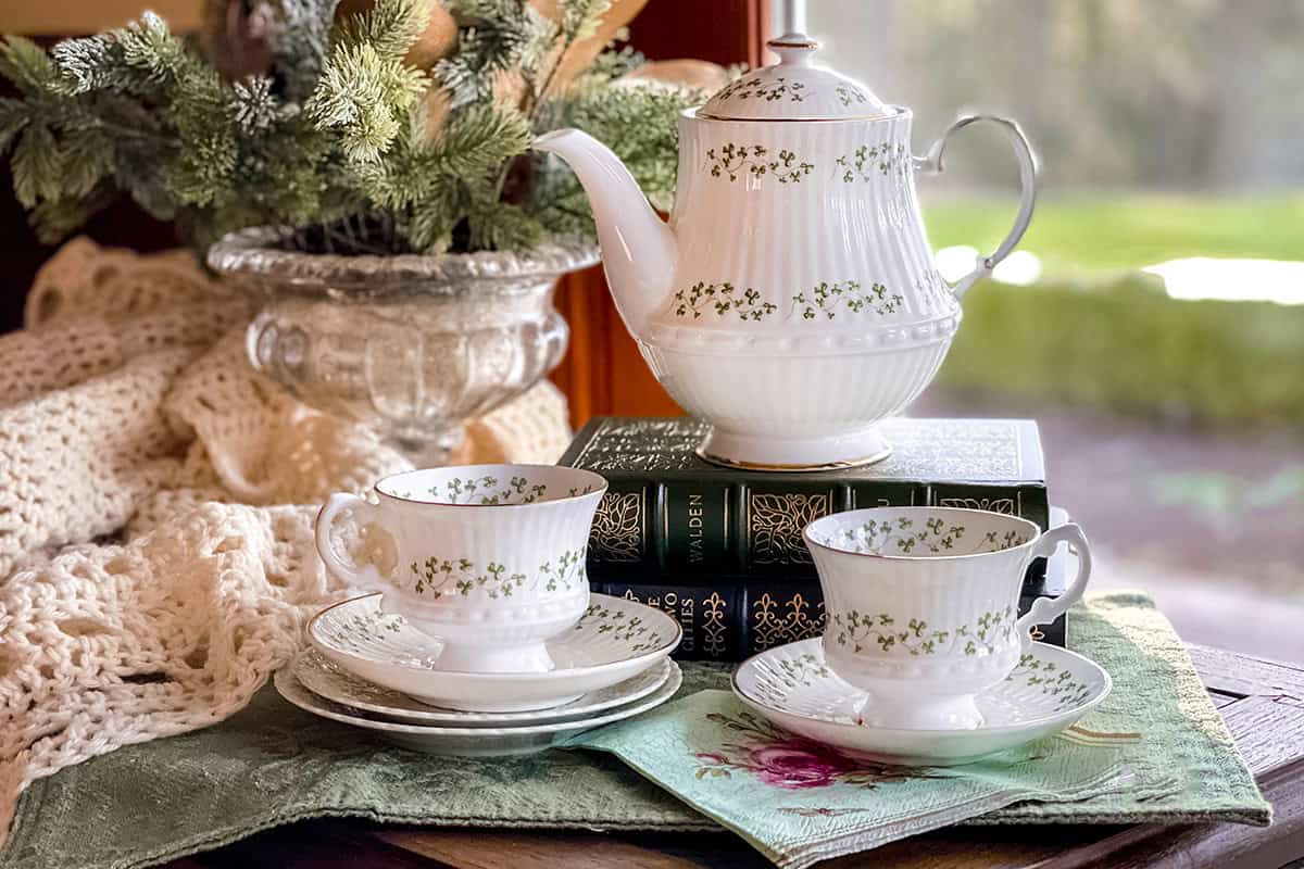 White china teapot and two teacups with green floral patterns evoke the charm of Irish Afternoon Tea, arranged on stacked books and a napkin, with a cozy knitted blanket and plant in the background.