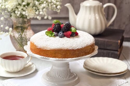 An Irish Tea Cake dusted with powdered sugar is elegantly topped with blackberries, raspberries, blueberries, and mint leaves on a pristine white cake stand. Nearby sit a teapot, a cup of tea, a stack of books, and a vase with white flowers.