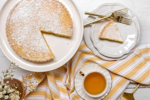 Horizontal top view of a sliced lemon custard tart with a wedge sitting on a nearby plate. Both the slice and the tart are dusted generously with powdered sugar, and a teacup nestled between contains an amber tea.