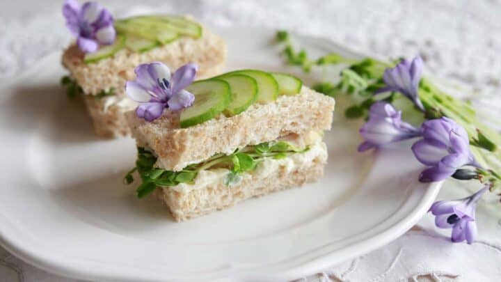 Two triangular cucumber sandwiches on a white plate, garnished with purple flowers. The sandwiches are layered with cream filling and microgreen leaves, placed on a delicate lace tablecloth.