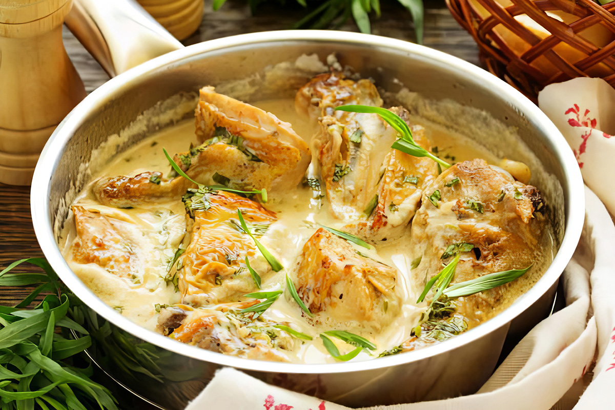 A stainless steel pan filled with Abraham Lincoln's Chicken Fricassee in a creamy sauce, garnished with fresh herbs, sits on a wooden table with a cloth and knife nearby.