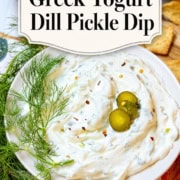 A plate of creamy dill pickle dip made with Greek yogurt, garnished with fresh dill and pickles. Crackers, extra pickles, and a towel complete the background. Text on image: "2 Minute Greek Yogurt Dill Pickle Dip.
