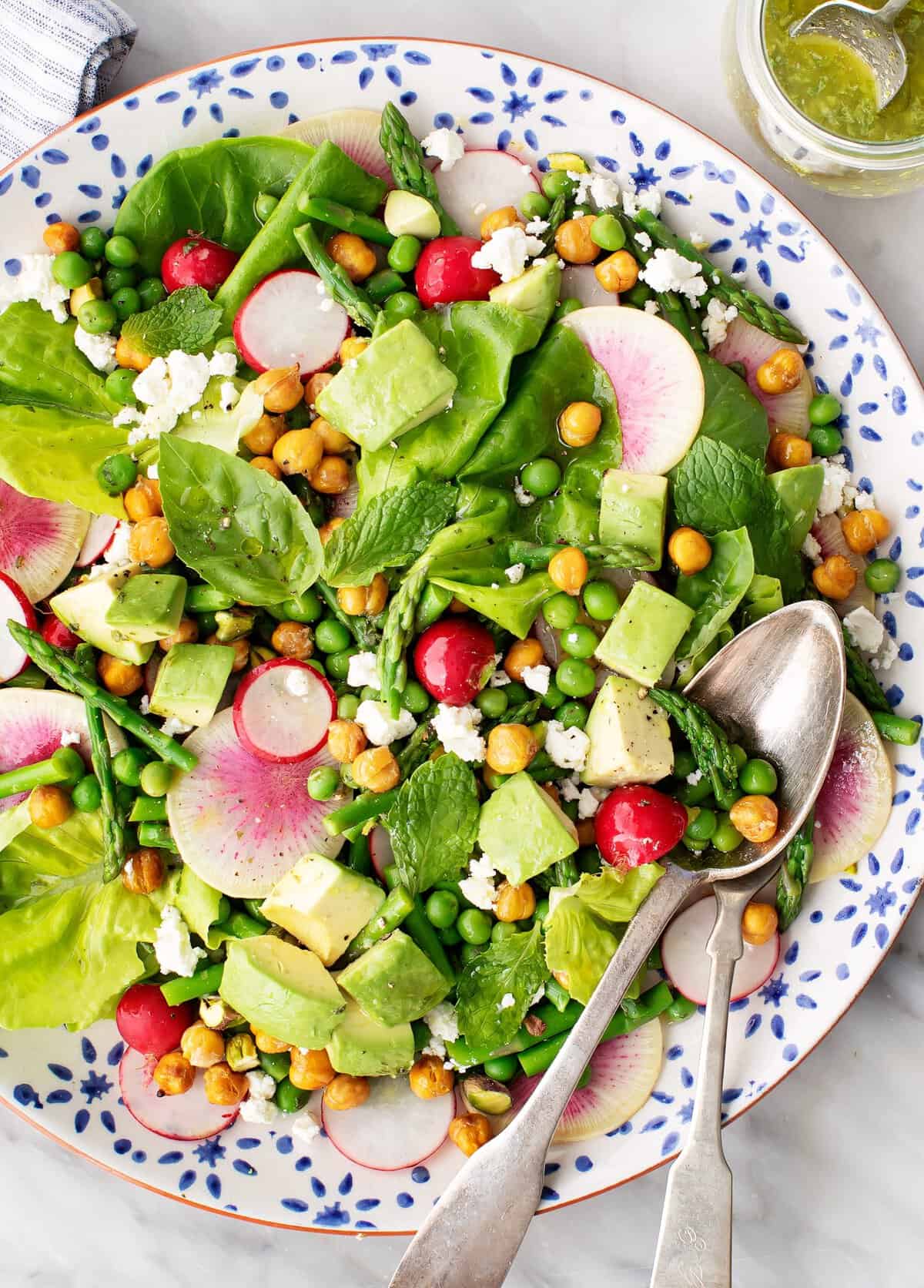 A vibrant salad on a decorative plate features leafy greens, avocado cubes, radish slices, peas, roasted chickpeas, and crumbled feta. A jar of dressing is beside the plate, and a spoon and fork rest on the salad.