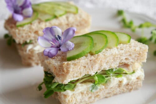 Two cucumber tea sandwiches with microgreens and herbed cream cheese, is topped with thinly sliced cucumbers and edible flowers.