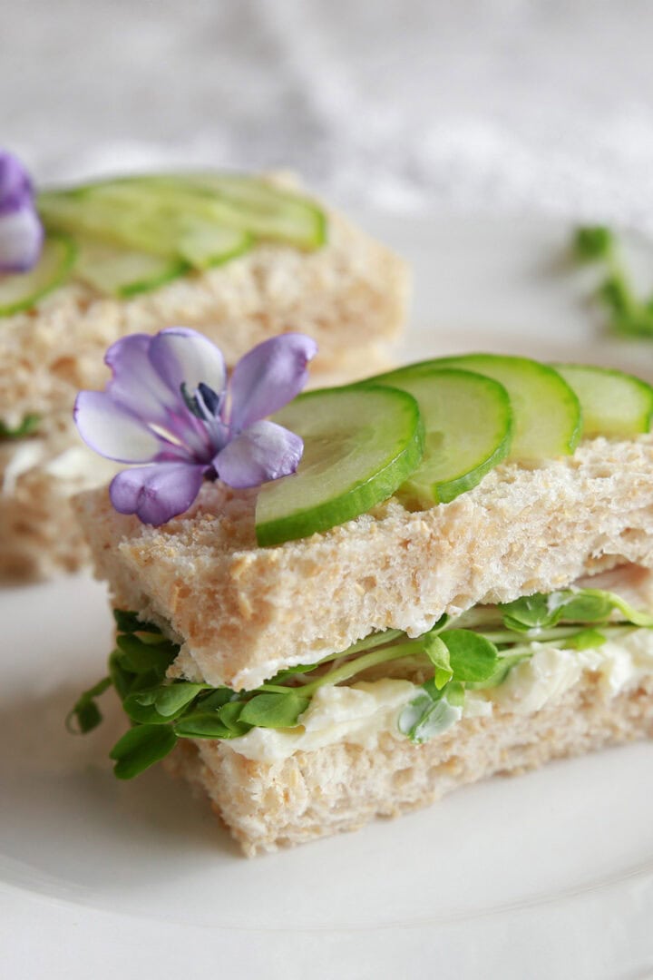Two cucumber tea sandwiches with microgreens and herbed cream cheese, is topped with thinly sliced cucumbers and edible flowers.
