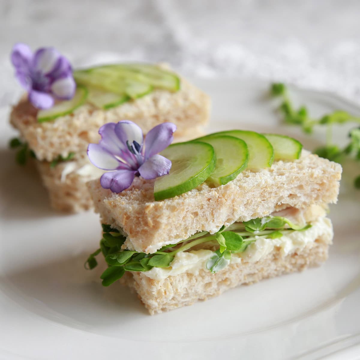 Two cucumber tea sandwiches with microgreens and herbed cream cheese, is topped with thinly sliced cucumbers and edible flowers.
