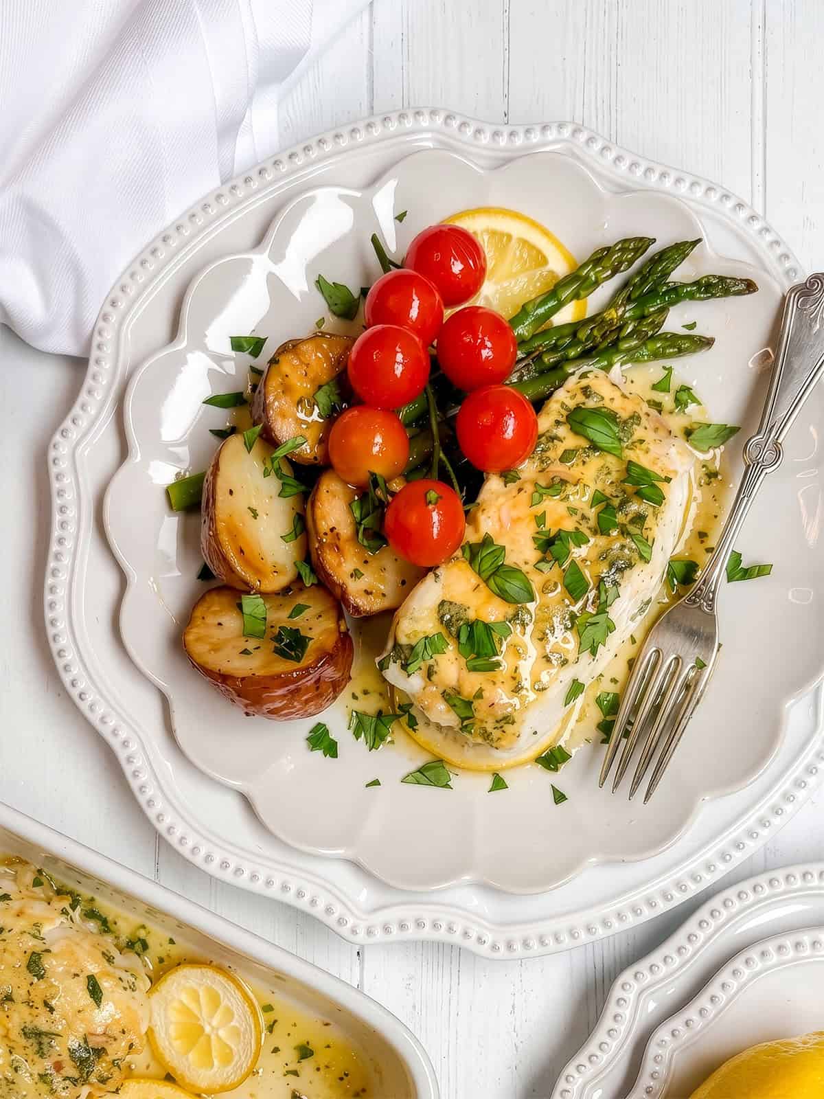 A plate with baked chicken breast in lemon herb sauce, roasted potatoes, asparagus, and cherry tomatoes, garnished with fresh herbs. A fork rests on the plate alongside a portion of flaky baked cod.