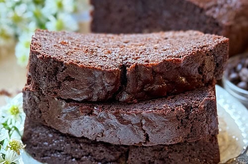 Side view of 3 slices of chocolate loaf cake, resting on a white plate, with small white flours laying nearby. The rest of the loaf is in the background.