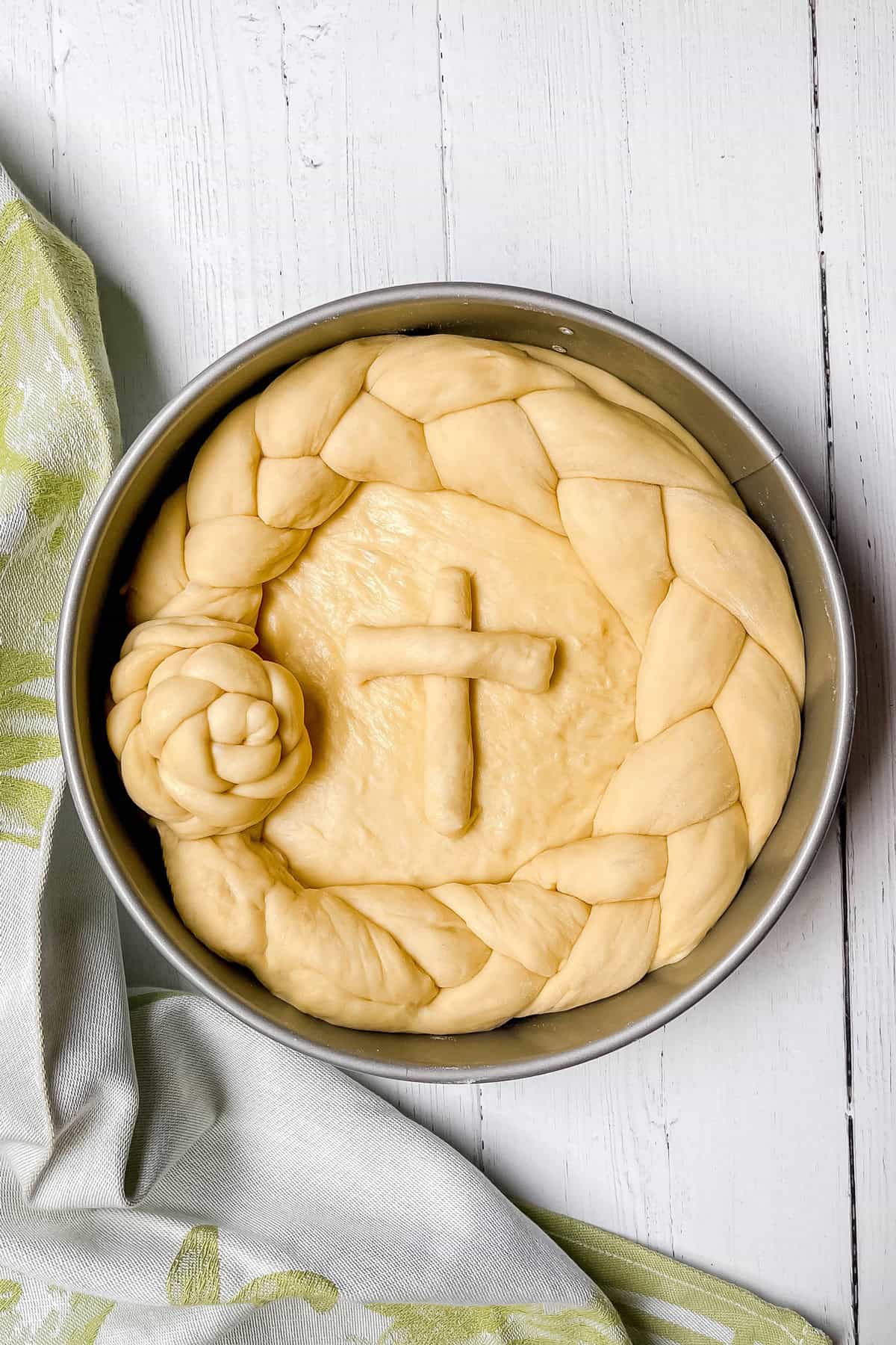 Unbaked round loaf of paska dough in a pan, decorated with a braided border and a cross on top, on a white wooden surface next to a light green cloth.