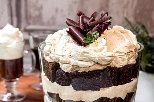 Side view of a layered Irish Coffee Trifle ready for serving in a glass trifle bowl, decorated with cream, chocolate curls, and fresh mint sprigs. Mugs of Irish Coffee is in the background.