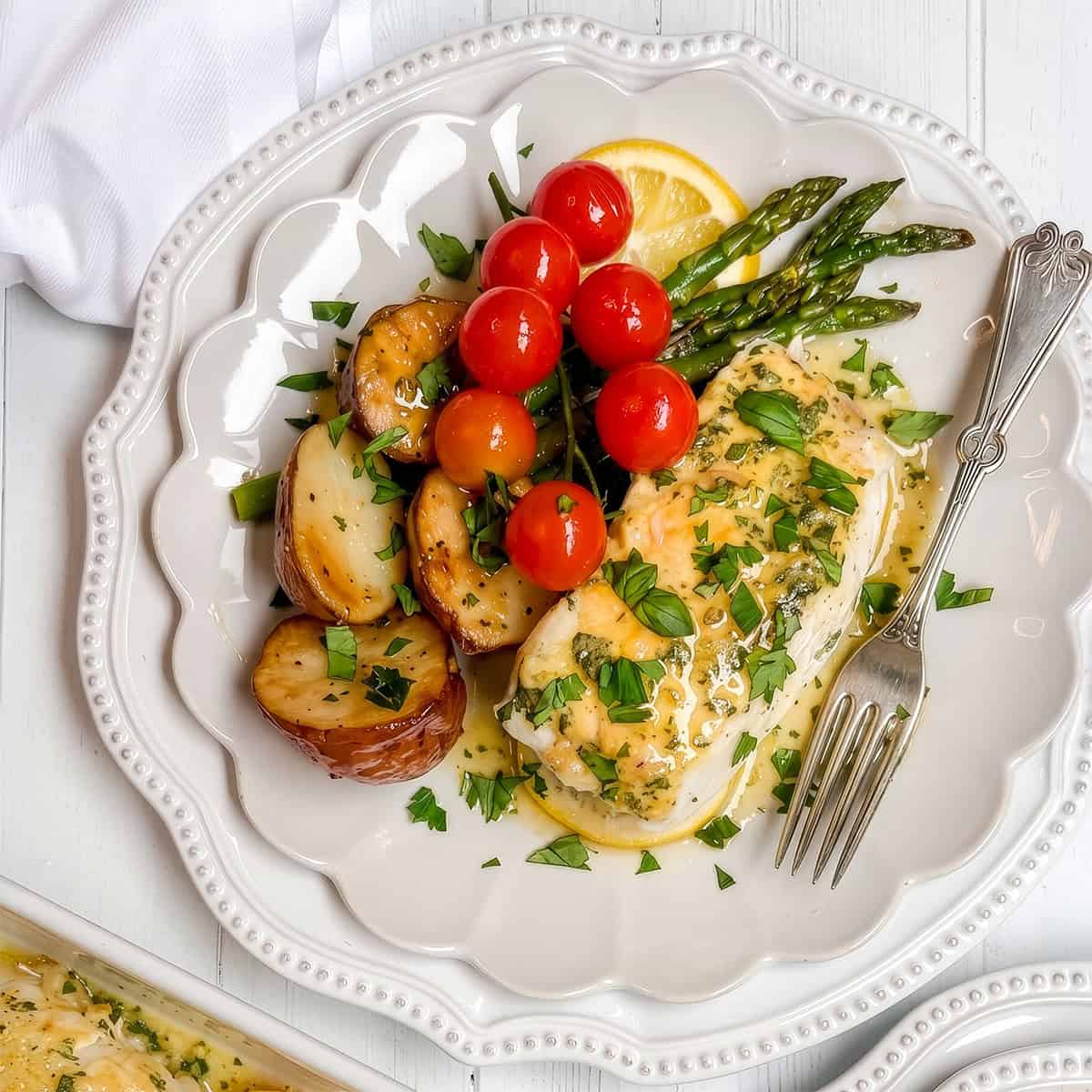 A plate with baked cod topped with herbs, roasted potatoes, asparagus, cherry tomatoes, and a slice of lemon, served with a fork on a white plate.