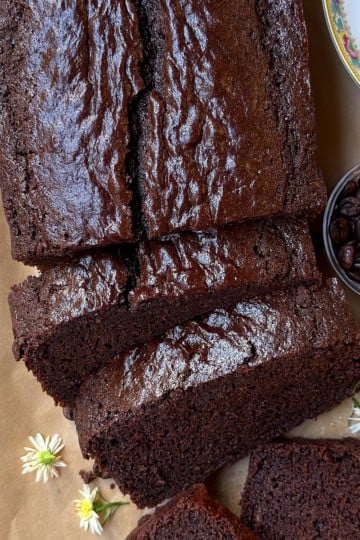 Partially sliced chocolate loaf cake on parchment paper with a serrated knife nearby.