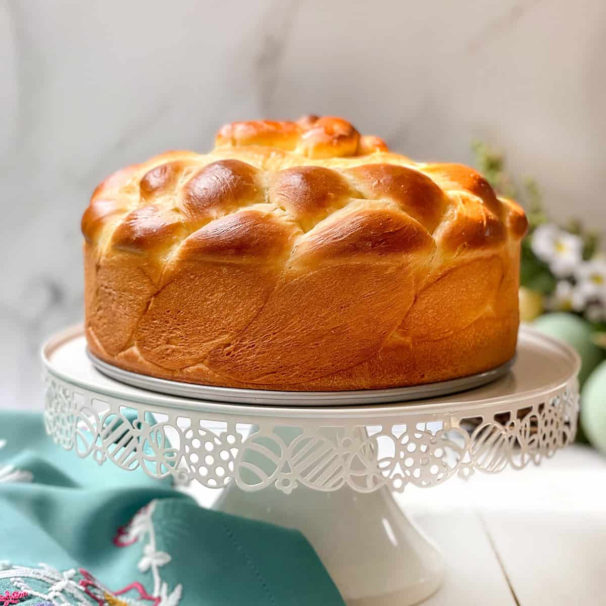 Side view of unsliced Easter Paska yeasted bread on a white spring stand with Easter eggs in the background.