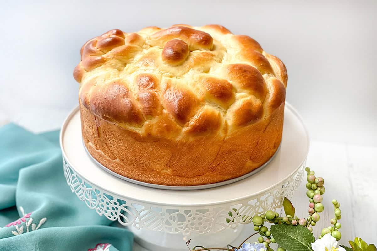 A round, golden-brown braided paska bread sits on a white decorative cake stand, with a teal cloth and flowers nearby.