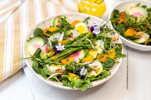 A fresh salad with asparagus, sliced radishes, cucumbers, carrots, leafy greens, and edible flowers is served in a white bowl. A yellow-patterned cloth and a carafe with yellow dressing are in the background.