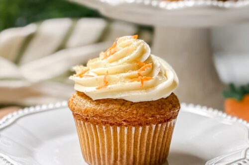 A carrot cake cupcake crowned with swirled cream cheese frosting and a sprinkle of orange zest rests on a white plate. More cupcakes are showcased in the background on a tiered stand, creating a light and airy setting with a soft-focus backdrop.