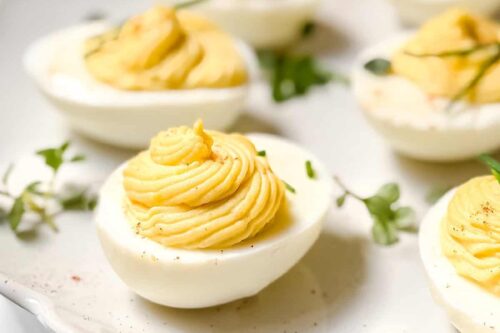 Close-up of a plate with deviled eggs, each topped with creamy yellow filling. This delightful deviled eggs recipe is garnished with small green herbs, neatly arranged on a light-colored plate, creating an appetizing presentation.