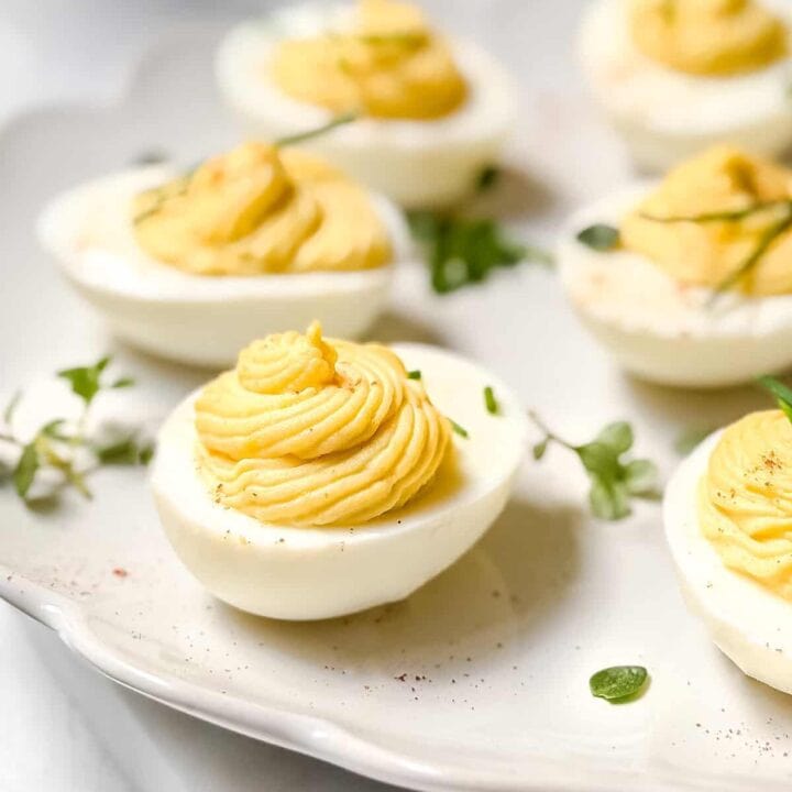Close-up of a plate with deviled eggs, each topped with creamy yellow filling. This delightful deviled eggs recipe is garnished with small green herbs, neatly arranged on a light-colored plate, creating an appetizing presentation.