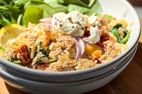 A bowl of orzo pasta with ground turkey skillet, sun-dried tomatoes, red onions, spinach, and a dollop of creamy sauce, served with fresh spinach leaves and a lemon wedge on the side.