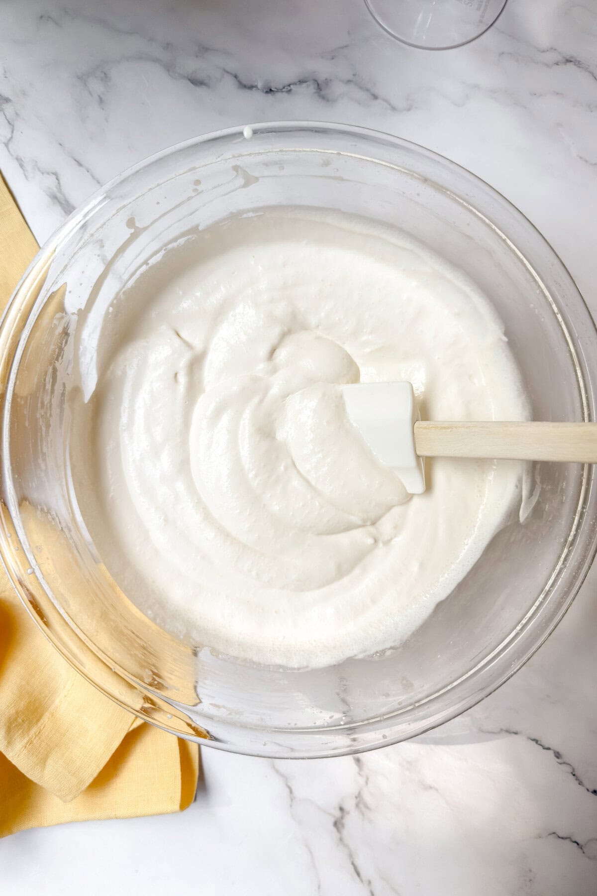 A bowl of smooth, creamy white batter is being mixed with a white spatula, ready to become a delightful dessert. The bowl sits on a marble surface, with a folded yellow cloth adding a pop of color beside it. Perfect for layering into an elegant trifle.