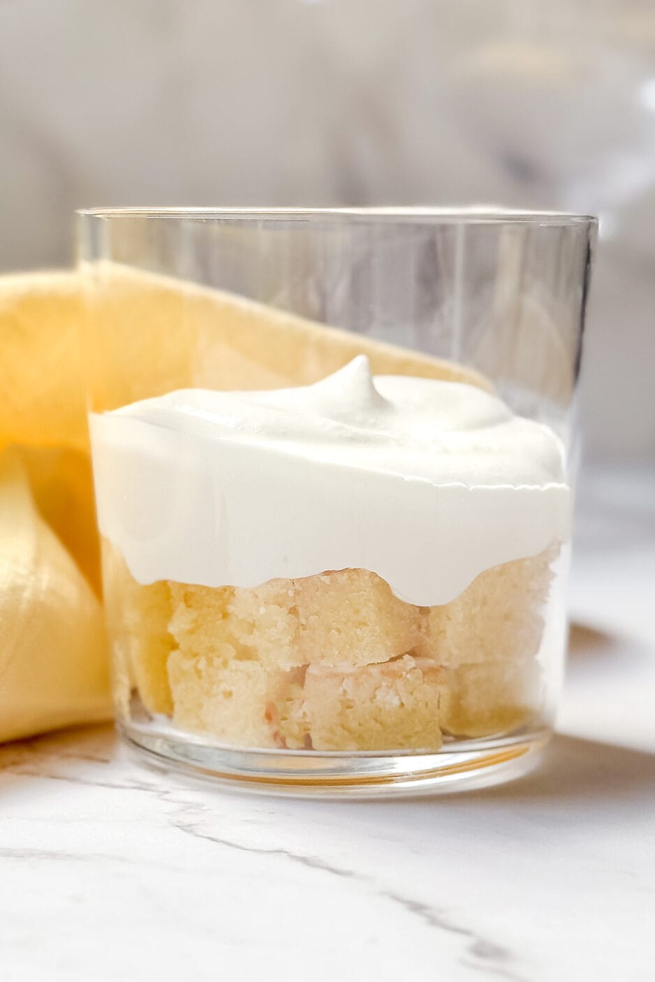 A glass dessert trifle cup brimming with layers of light cake and crowned with smooth whipped cream sits elegantly on a marble surface, complemented by a yellow napkin in the background.