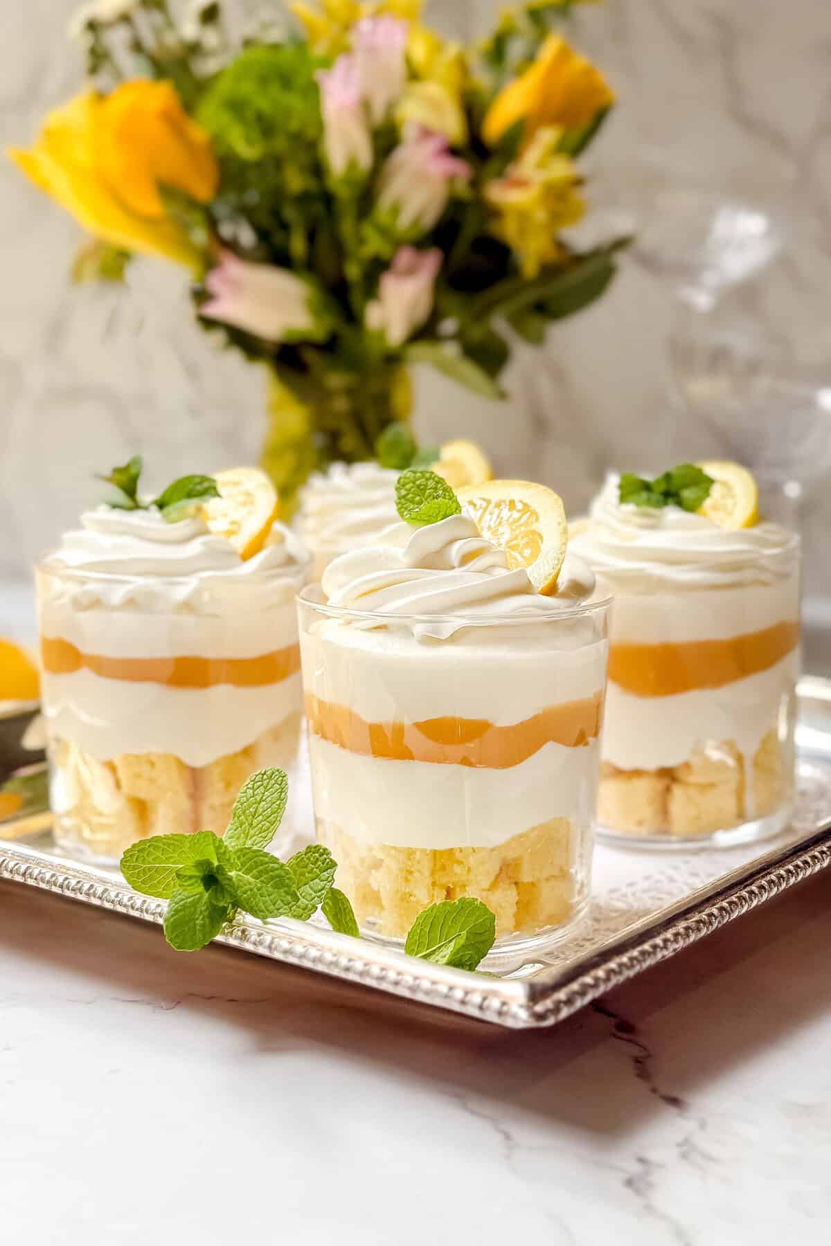 Elegant dessert trifle cups layered with sponge cake, lemon curd, and whipped cream, garnished with lemon slices and mint leaves. Arranged on a silver tray with a vase of yellow and white flowers in the background.