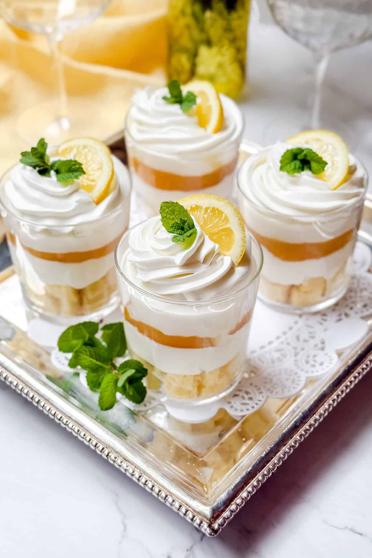 Four layered dessert trifle cups with whipped cream, lemon slices, and mint leaves rest on a silver tray. The base of cake or biscuit layers adds depth to each cup, set elegantly on a lace doily. A blurred background reveals glasses and a green bottle.