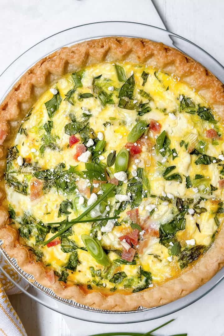 A baked asparagus quiche with a golden crust, filled with eggs, spinach, diced tomatoes, and green onions, garnished with herbs and crumbled cheese, displayed on a white surface.