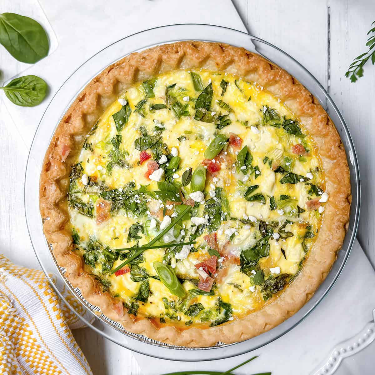 A baked asparagus quiche with a golden crust, filled with eggs, spinach, diced tomatoes, and green onions, garnished with herbs and crumbled cheese, displayed on a white surface.