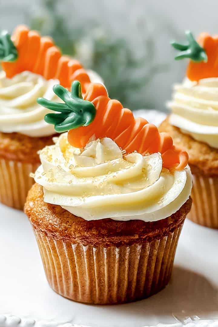 Three carrot cake cupcakes with white frosting are topped with decorative orange and green carrot-shaped toppers, displayed on a white plate.
