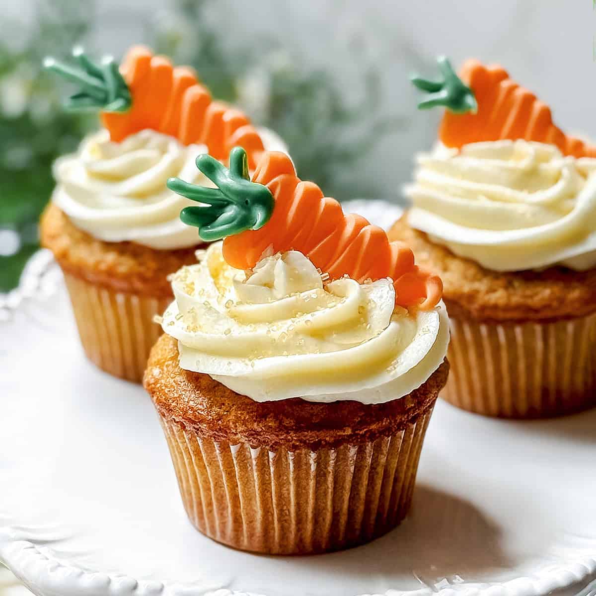 Three carrot cake cupcakes with white frosting are topped with decorative orange and green carrot-shaped toppers, displayed on a white plate.