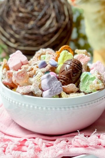 A white bowl filled with colorful Easter Snack Mix—cereal, heart-shaped marshmallows, and a chocolate egg—rests on a pink napkin, with a nest and giraffe figurine in the background.